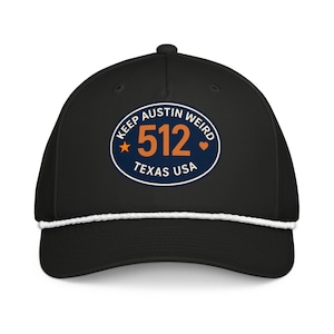 Puede incluir: Gorra de béisbol negra con detalle de cuerda blanca y un parche circular. El parche dice "KEEP AUSTIN WEIRD 512 TEXAS USA" en blanco, naranja y azul. Una estrella y un corazón acentúan el texto. La gorra tiene una visera curva.