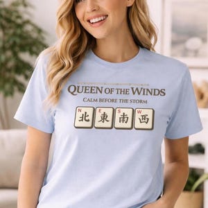 Op de afbeelding: Lichtblauw T-shirt met de tekst "QUEEN OF THE WINDS" en "CALM BEFORE THE STORM" in bruin. Daaronder vier tegels met Chinese karakters en de letters N, E, S en W.