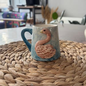 Op de afbeelding: Een keramische mok met een blauw handvat en een lichtblauwe body. De mok heeft een reliëf van een roze zwaan met gedetailleerde veren. De achtergrond heeft een aquareleffect. De mok staat op een geweven placemat.