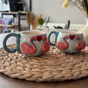 Op de afbeelding: Twee keramische mokken met blauwe handvatten en een lichtblauwe achtergrond. Elke mok heeft twee roze zwanen die tegenover elkaar staan met een rood hart ertussen. De mokken staan op een geweven placemat.