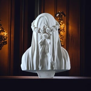 May include: A white bust sculpture of a veiled figure, adorned with a floral crown and a draped veil. The sculpture has a smooth finish and is set against a dark, wooden background. The bust is a decorative piece.