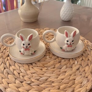 Puede incluir: Dos tazas de té y platillos de cerámica blanca con figuras de conejos. Los conejos tienen orejas rosas y pajaritas. Los platillos tienen un borde dorado. Las tazas y los platillos están sobre una estera tejida.