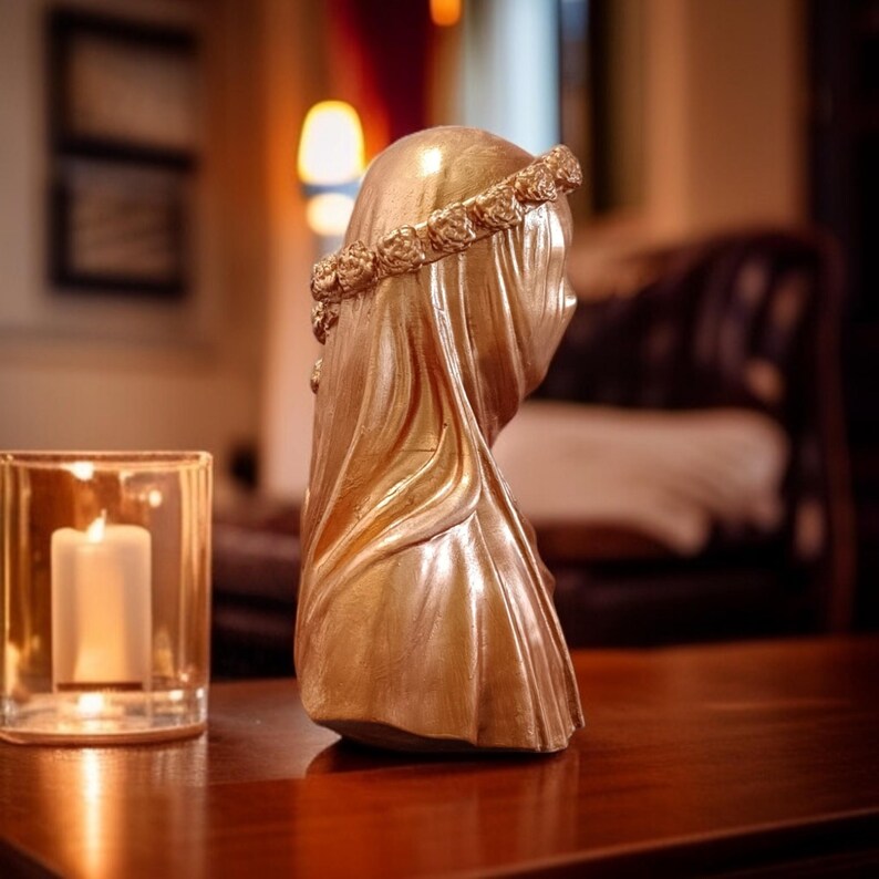 May include: A gold-colored bust of a veiled figure, adorned with a floral crown. The sculpture is placed on a wooden surface, next to a lit candle in a glass holder. The bust is a decorative item.
