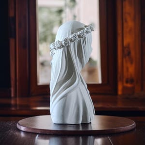 May include: A white bust of a veiled figure, adorned with a floral crown. The sculpture is set on a round, dark wooden base, with a blurred background of a window and wooden paneling.