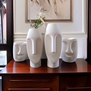 Puede incluir: Cuatro jarrones blancos de cerámica con forma de rostro de diferentes tamaños se exhiben sobre una superficie de madera oscura. Los jarrones tienen un diseño minimalista, con rostros alargados con narices prominentes y rasgos sutiles. Decoración del hogar.