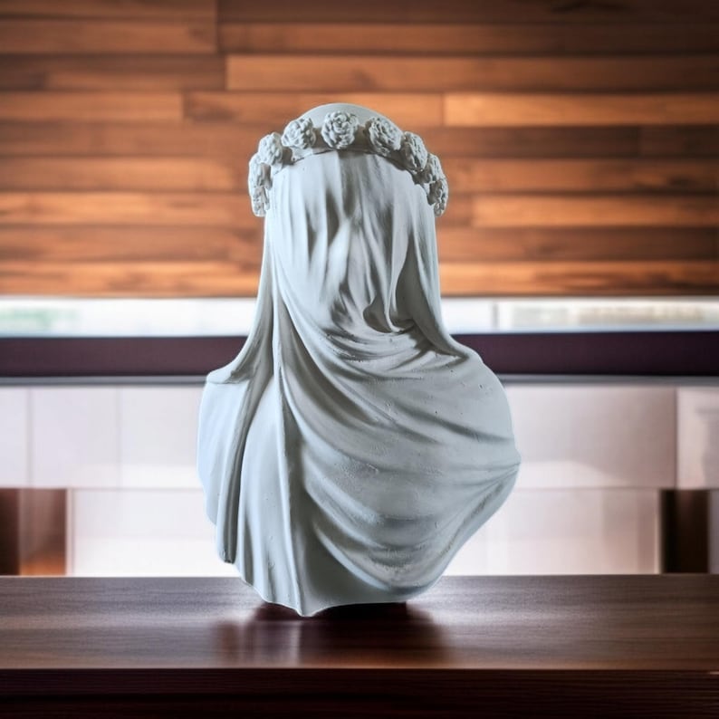 May include: A white bust sculpture of a veiled figure, adorned with a floral crown. The draped fabric cascades down, obscuring the face. The sculpture is set against a wooden background.