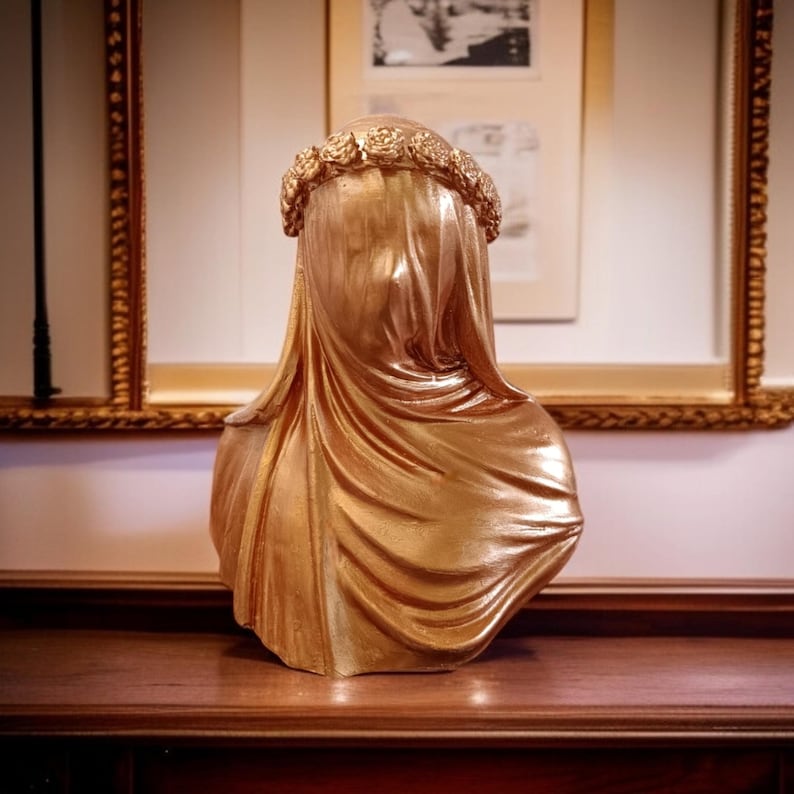 May include: A gold-colored bust sculpture of a veiled figure, adorned with a floral crown. The sculpture has a draped veil and is displayed on a wooden surface, with framed artwork in the background.