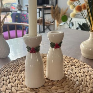 Puede incluir: Dos candelabros de cerámica blanquecina con un diseño de acebo y bayas. Uno sostiene una vela blanca. Los candelabros están sobre una estera tejida, con jarrones y decoración en el fondo.