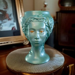 Könnte beinhalten: Türkisfarbene Büste einer Frau mit orangefarbenen Augen und lockigem Haar. Die Skulptur hat ein klassisches Design und steht auf einer runden, grauen, gewebten Oberfläche. Die Büste besteht aus glänzendem Material.