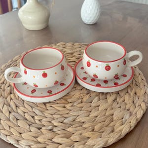 May include: Two white ceramic teacups and saucers with a red rose pattern. The cups have handles and are placed on matching saucers. The rims and edges are outlined in red. The set is on a woven placemat.
