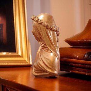May include: A gold-colored bust sculpture of a veiled figure, adorned with a floral crown. The sculpture has a draped fabric design and is set against a blurred background.