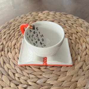 May include: White ceramic teacup and saucer set. The cup has a red pencil-shaped handle and scattered black letters inside. The saucer is designed to look like an open book with an orange border.