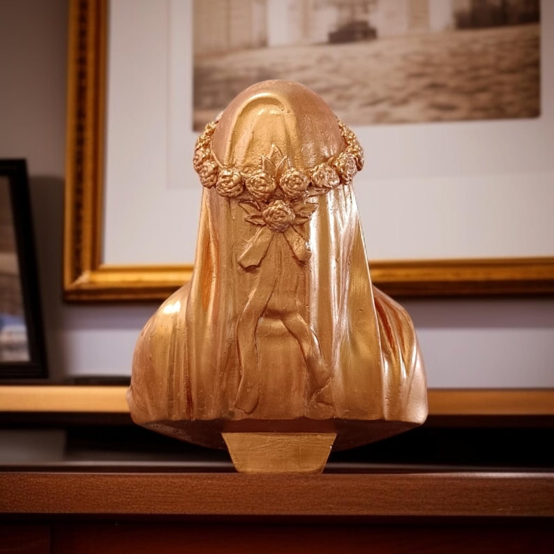 May include: A gold-colored bust sculpture of a veiled figure, adorned with a floral crown. The sculpture features detailed drapery and a bow design on the veil. The bust is displayed on a wooden surface, with framed artwork in the background.