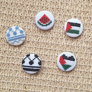 Puede incluir: Una colección de cinco broches redondos bordados. Los broches presentan varios diseños, incluyendo una rodaja de sandía, patrones geométricos y la bandera palestina. Los broches son blancos con costuras coloridas.