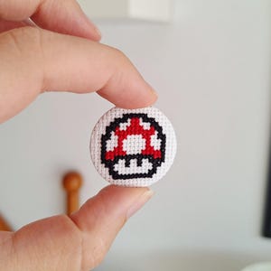 Op de afbeelding: Een kleine, ronde, witte kruissteekknop met een rood en wit paddenstoelontwerp en zwarte contouren. De knop wordt tussen twee vingers gehouden. Het paddenstoelontwerp is gepixeld, vergelijkbaar met een videogame.