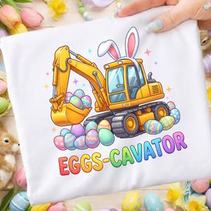 May include: White t-shirt featuring a yellow excavator wearing bunny ears, scooping up colorful Easter eggs. The text "EGGS-CAVATOR" is printed in rainbow colors below the image. A fun, festive design.