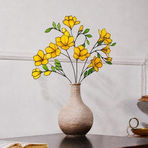 May include: A stained glass arrangement of bright yellow flowers with green leaves on thin dark stems, displayed in a textured, light brown ceramic vase with a rounded base and a narrow neck, creating a decorative botanical sculpture.