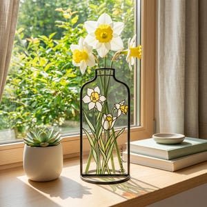 Op de afbeelding: Een heldere glazen vaas met een zwart metalen frame, met witte en gele narcissen. De vaas heeft een decoratief narcissenontwerp. Een kleine vetplant in een witte pot staat in de buurt. De vaas is ongeveer 25 cm hoog.