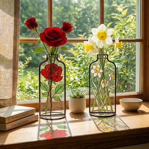 Op de afbeelding: Twee decoratieve glazen vazen met zwarte metalen frames, een met rode rozen en de andere met witte en gele narcissen. De vazen staan op een houten vensterbank, met een kleine vetplant en een kom.