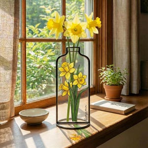 Op de afbeelding: Een heldere glazen vaas met een zwart metalen frame, met gele narcissen. De vaas heeft een geschilderd narcissenontwerp en de naam "Benjamin". De vaas staat op een houten vensterbank met een kleine kom en een potplant.