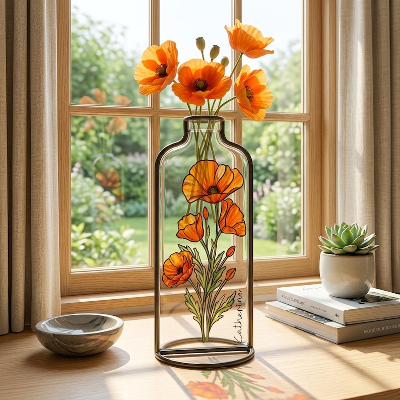 Op de afbeelding: Een heldere glazen vaas met een zwart metalen frame, met een geschilderd klaproosontwerp en verse oranje klaprozen. De vaas staat op een houten oppervlak bij een raam. De naam "Catherine" staat op de vaas.