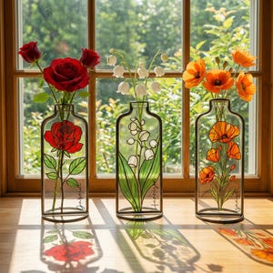 Op de afbeelding: Drie decoratieve glazen vazen met zwarte metalen frames, elk met verschillende bloemen. De eerste vaas heeft een rood rozenontwerp en echte rode rozen. De tweede vaas heeft een lelietjes-van-dalenontwerp en echte witte bloemen. De derde vaas heeft een oranje klaprozenontwerp en echte oranje klaprozen.
