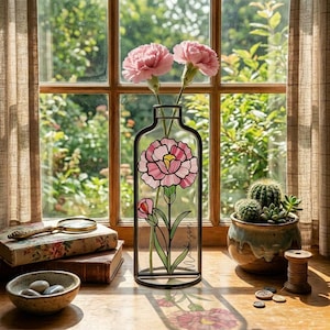 Op de afbeelding: Een heldere glazen vaas met een zwart metalen frame, met een roze bloemenontwerp en de naam "Madison". Twee roze anjers staan in de vaas. Een kleine cactus in een keramische pot staat op een houten oppervlak bij een raam.