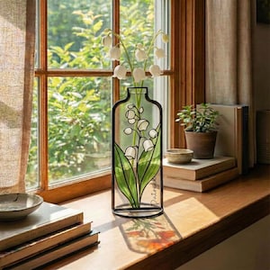 Op de afbeelding: Een decoratieve zwarte metalen vaas met een glas-in-lood ontwerp van lelietjes-van-dalen bloemen. De vaas bevat witte bloemen en groene bladeren. De vaas staat op een houten vensterbank bij een raam.