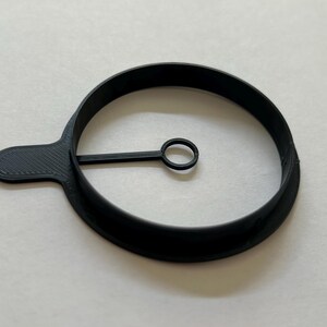 Può includere: Anello per uova in plastica nera con manico. L'anello ha un cerchio più piccolo al centro.