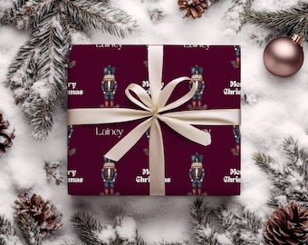 Pode incluir: Um presente embrulhado com um fundo bordô com um padrão de soldados quebra-nozes e as palavras "Merry Christmas" e "Lainey" em branco. O presente é amarrado com uma fita e laço creme, e é cercado por galhos de pinheiro e neve.