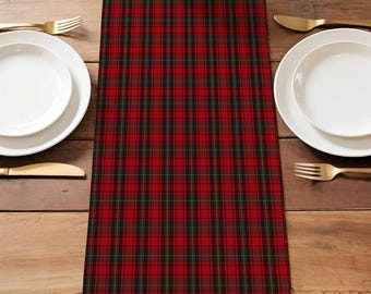May include: A red and black plaid table runner with green accents. The runner is centered on a wooden table, with white plates, gold cutlery, and a woven basket visible. The plaid pattern is a classic design.