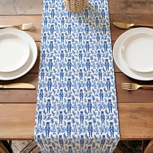 May include: A white table runner with a repeating pattern of blue nutcracker soldiers and leafy sprigs. The runner is set on a wooden table with white plates, gold cutlery, and a woven basket.