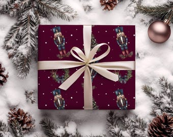 Pode incluir: Uma caixa de presente embrulhada com um fundo bordô com um padrão repetido de quebra-nozes, árvores de Natal e coroas. A caixa é amarrada com uma fita e laço creme. A cena é definida em uma superfície nevada com agulhas de pinheiro e pinhas.