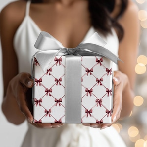 May include: A wrapped gift box with a white background and a pattern of burgundy bows. The box is tied with a silver ribbon and bow. The gift is held by a person wearing a white dress.