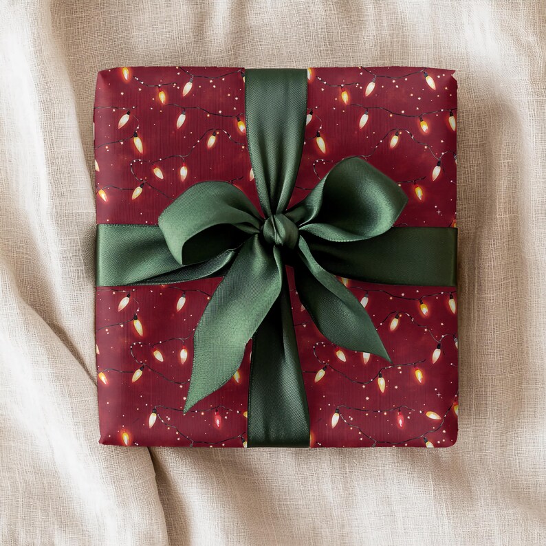 May include: A wrapped gift with a dark red background featuring a pattern of white lights. The gift is tied with a dark green satin ribbon and bow. The gift is presented on a neutral-colored fabric surface.