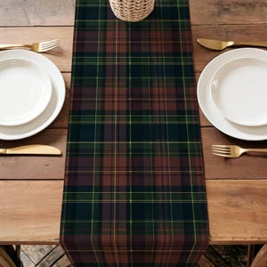 May include: A tartan table runner in brown, green, and navy blue, with yellow lines. The runner is on a wooden table, with white plates, gold cutlery, and a woven basket. The table setting is complete.