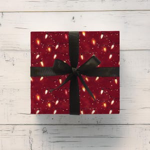 May include: A wrapped gift box with a festive design. The wrapping paper is a deep red color and features a pattern of white, yellow, and orange Christmas lights. A black ribbon is tied around the box in a bow.