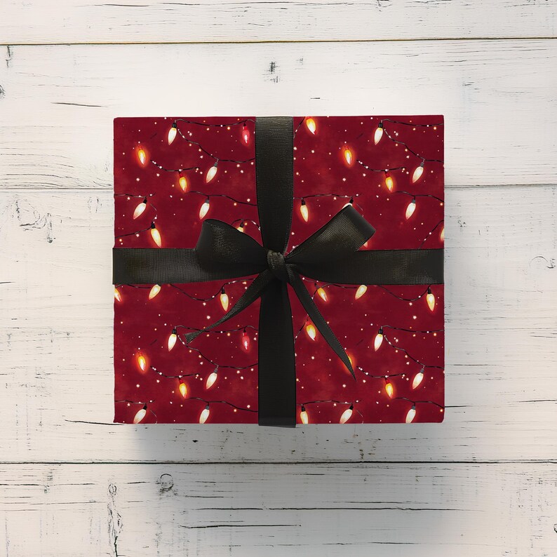 May include: A wrapped gift box with a festive design. The wrapping paper is a deep red color and features a pattern of white, yellow, and orange Christmas lights. A black ribbon is tied around the box in a bow.