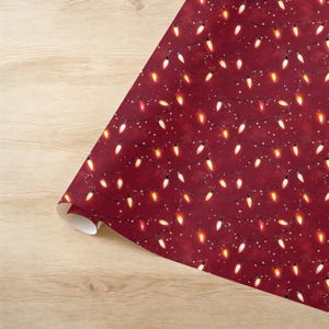 May include: A roll of wrapping paper with a festive design. The paper features a deep red background adorned with strings of Christmas lights in various colors, including white, yellow, and red. Small white dots add to the holiday theme.