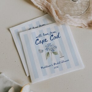 Könnte beinhalten: Zwei quadratische Papierservietten mit hellblauen vertikalen Streifen. Der Text lautet with love from Cape Cod, Maddison's Bridal Shower 2026. Die Illustrationen zeigen eine blaue Hortensienblüte, ein kleines Segelboot und eine Austernschale.