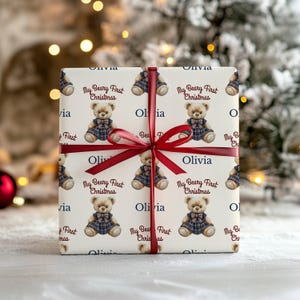 May include: A wrapped gift with a repeating pattern of teddy bears in blue plaid dresses, with the text "My Beary First Christmas" and the name "Olivia". The gift is tied with a burgundy ribbon.
