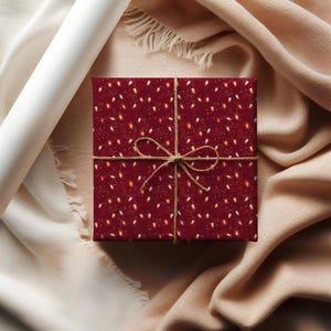 May include: A wrapped gift box with a festive pattern of white and gold lights on a deep red background. The box is tied with twine and sits on a beige fabric surface. A roll of white paper and a beige fabric are in the background.