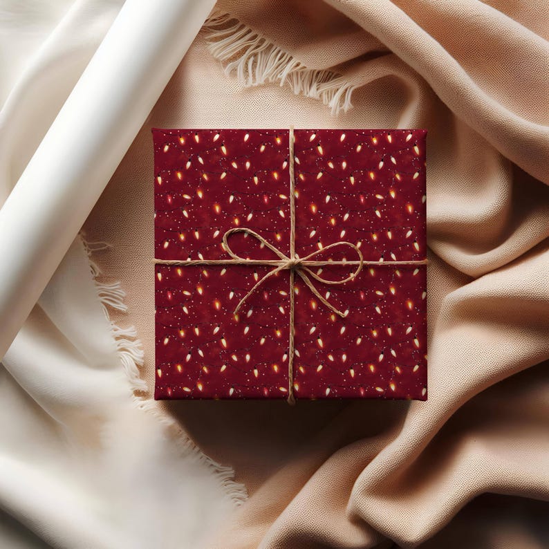 May include: A wrapped gift box with a festive pattern of white and gold lights on a deep red background. The box is tied with twine and sits on a beige fabric surface. A roll of white paper and a beige fabric are in the background.