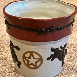 May include: A ceramic planter with a white body and a wide red rim. The rim features a raised black barbed wire design. The white exterior displays a repeating pattern of brown embossed reindeer silhouettes and a central brown star inside a circle.