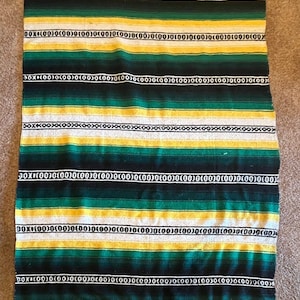 May include: A rectangular woven blanket featuring horizontal stripes in shades of dark green, yellow, and white. The design includes repeating black and white geometric patterns of circles and lines across the fabric, finished with a fringed edge.
