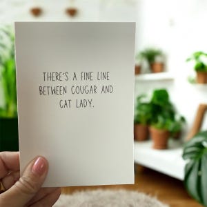 May include: A white greeting card with black text that reads "There's a fine line between cougar and cat lady." The card is held by a person. The background includes plants in pots.