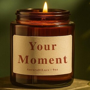May include: A lit candle in a brown glass jar with a white label that reads "Your Moment". The candle is 255g and sits on a green surface. The flame is bright and the background is a dark green.