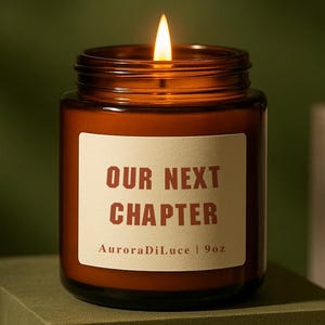 以下が含まれることがあります： 「OUR NEXT CHAPTER」と「AuroraDiLuce | 9oz」と書かれた白いラベルが付いた、ガラス瓶に入った、火の灯った琥珀色のキャンドル。キャンドルは緑色のブロックの上に置かれています。