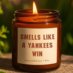 Pode incluir: Uma vela acesa em um frasco de vidro marrom com um rótulo branco que diz "Smells Like A Yankees Win". A vela tem 255g. O fundo é folhagem verde desfocada e uma superfície de pedra.
