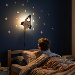 Könnte beinhalten: Eine holzfarbene Wandlampe in Raketenform mit blau-weißem Design beleuchtet eine dunkelblaue Wand, die mit sternförmigen Ausschnitten verziert ist. Die Lampe ist eingesteckt und strahlt ein warmes Licht aus, wodurch eine gemütliche Atmosphäre in einem Kinderzimmer entsteht.